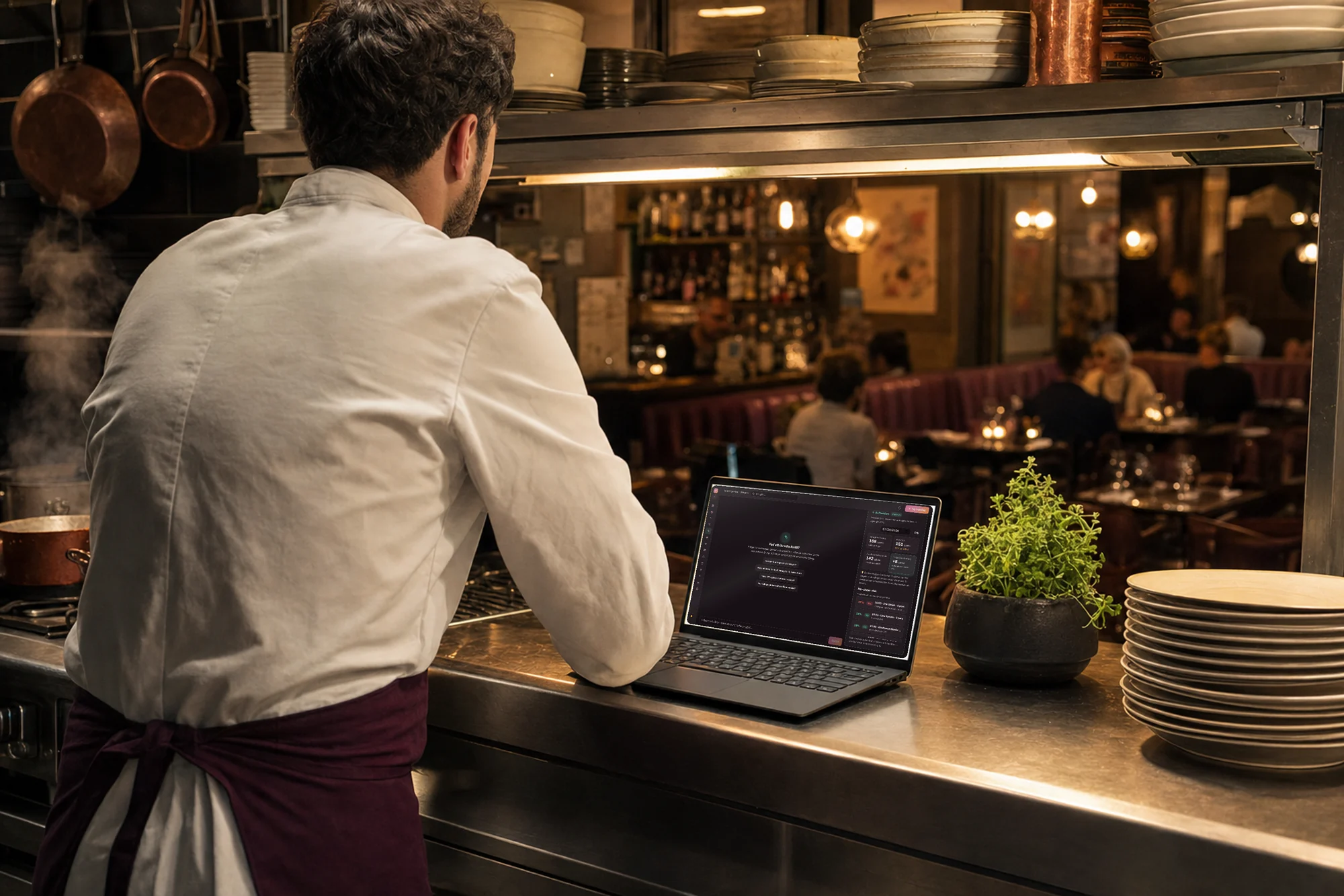 Kock vid kökspasset med Goboblo AI på laptopen — frågan Hur ser kvällen ut? besvarad med kvällens bokningar och no-show-risk, matsalen i bakgrunden