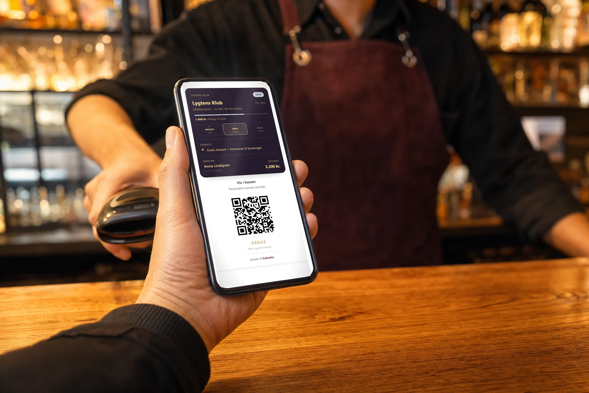 Gæstens hånd viser medlemskortet på telefonen ved kassen — Lygtens Klub på Sølv med QR-kode, personale med scanner klar i baggrunden