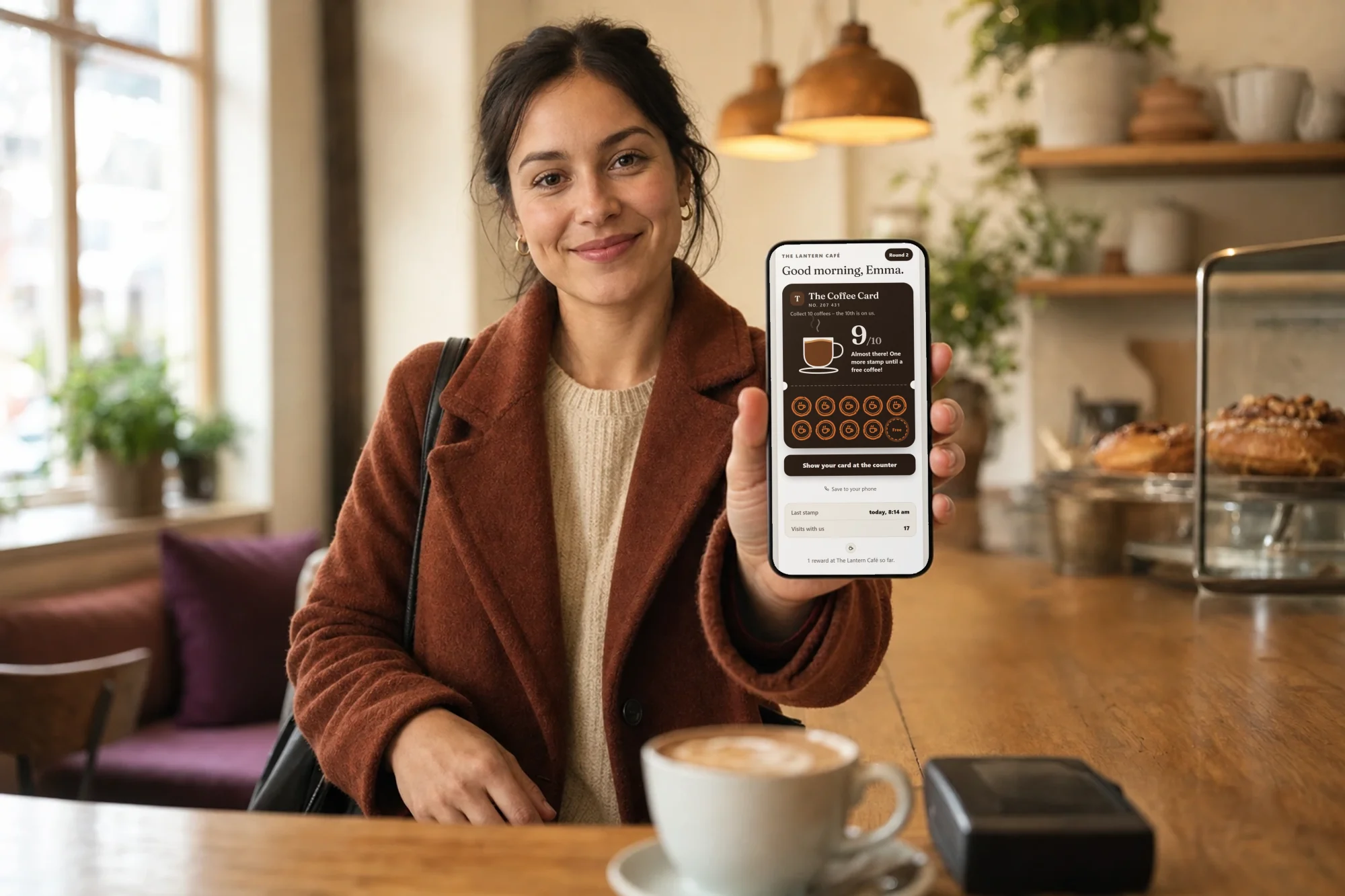 A guest in a café holds up her phone to the register. The screen shows the coffee stamp card with 9 of 10 stamps and the words “Almost there! One more stamp until a free coffee!”
